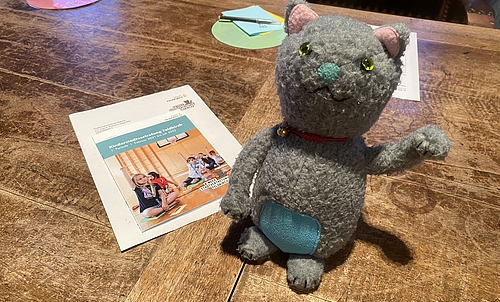 Das Maskottchen der Kinderstadtvertretung, die Katzenturm-Katze sitzt auf einem Tisch. Daneben liegt ein Folder mit Informationen zur Kinderstadtvertretung.