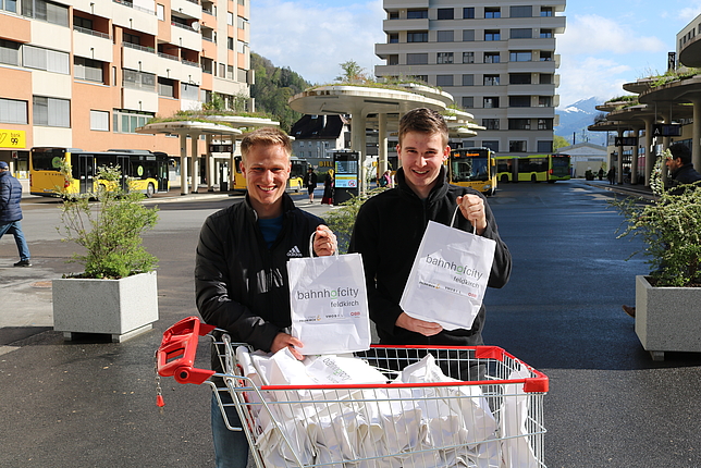 Zwei Zivildiener stehen auf dem Bahnhofsvorplatz vor einem Einkaufswagen. Der Einkaufswagen ist mit Papiersäcken gefüllt. Die zwei Jungs halten je einen Papiersack mit dem Schriftzug "Bahnhofcity" in den Händen.