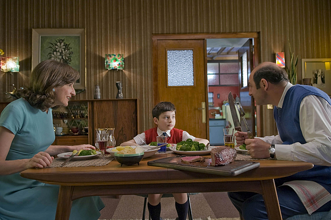 Ein kleiner Junge sitzt zwischen seiner Mutter und seinem Vater am Esstisch. Der Junge und seine Mutter schauen den Vater an.