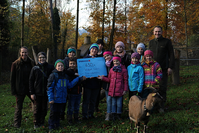 Kinder halten einen Scheck für den Wildpark in den Händen. Zwei Mitarbeiter:innen des Wildpark und eine Ziege stehen bei ihnen.