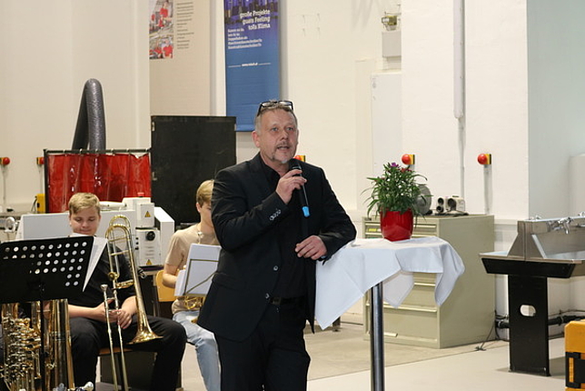 Direktor Hannes Mäser lehnt sich mit einem Arm auf einen Stehtisch, in der anderen Hand hält er ein Mikrofon, in das er spricht. Im Hintergrund sitzen zwei Jungen mit ihren Blasinstrumentn.