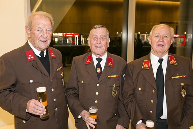 Drei ältere Feuerwehrmänner stehen nebeneinander, jeder hält ein Glas Bier in einer Hand