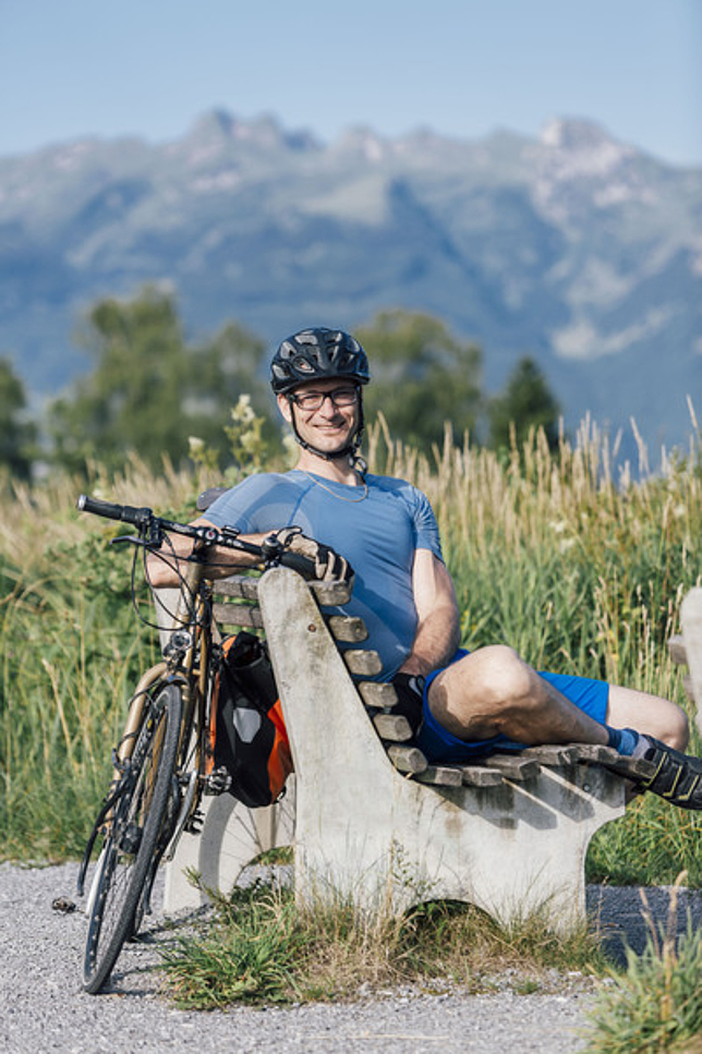 Ein Mann sitzt mit einem Fahrradhelm auf einer Parkbank, an die Bank gelehnt steht das Fahrrad.