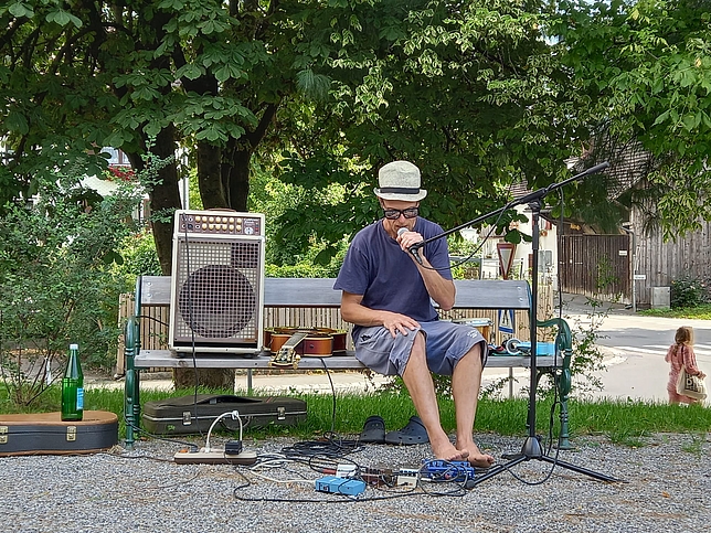 Ein Mann sitzt auf einer Holzbank, neben im liegt eine Gitarre und ein Lautsprecher auf der Bank. Er hat ein Mikrofon vor dem Mund, das er mit einer Hand hält. Mit den Füßen betätigt er ein Fußpedal.