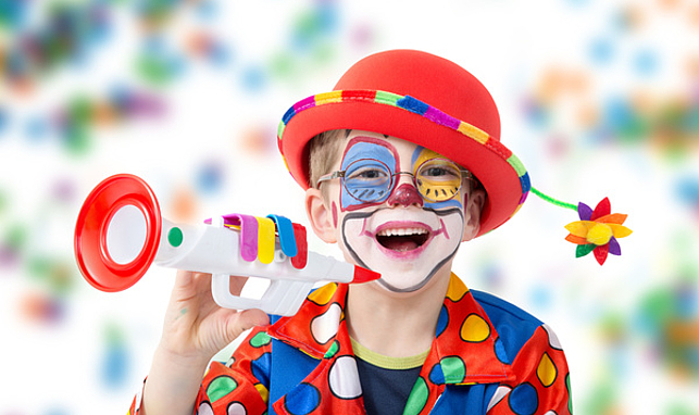 Ein kleiner Junge mit Brille ist als Clown verkleidet und hält in einer Hand eine Spielzeug-Trompete. 