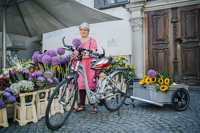 Eine Dame steht mit ihrem Fahrrad neben einem Blumenverkaufsstand.