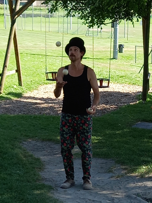 Ein Mann steht auf einem Spielplatz und jongliert mit drei Bällen. Er trägt einen Hut.