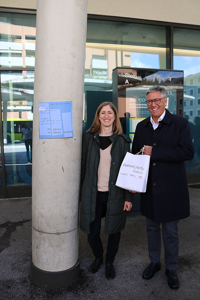 Bürgermeister Matt und Veronika Schubert stehen am Bahnhof neben einer Beton-Säule. Bürgermeister Matt hält einen Papiersack mit dem Schriftzug "Bahnhofcity" in den Händen. Auf der Säule ist ein blaues Viereck mit Text angebracht.