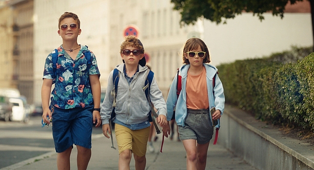 Zwei Jungen und ein Mädchen laufen auf einem Gehsteig. Alle tragen Sonnenbrillen, haben kurze Hosen an und haben einen Rucksack auf dem Rücken. 