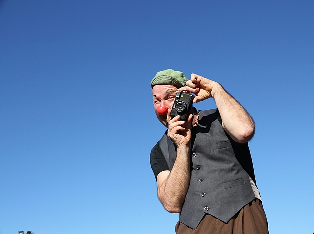 Ein Mann hält eine Kamera in den Händen und hält diese an sein Gesicht. Er trägt eine rote Clownsnase.