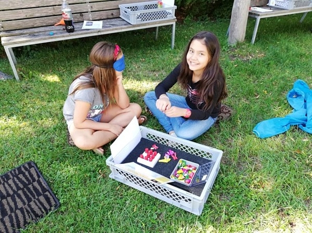 Zwei Mädchen sitzen mit einer Kiste voller Süßigkeiten in einer Wiese, eines der Mädchen hat die Augen verbunden. 