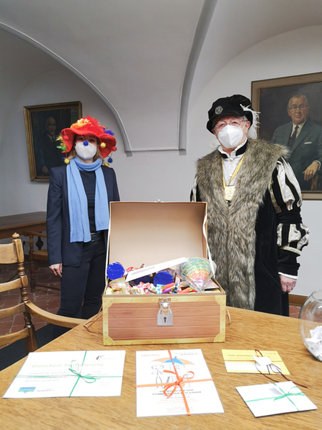 Stadträtin Gudrun Petz-Bechter und Graf Rudolf von Montfort stehen verkleidet vor einem Tisch mit Gutscheinen und Faschingsdekoartikeln.