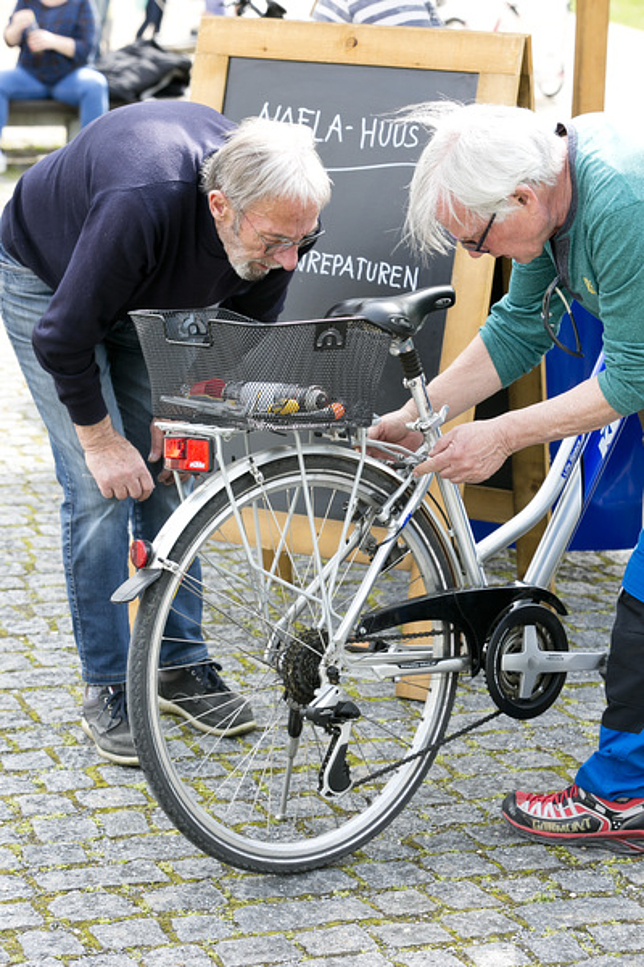 Ein älterer Mann steht neben einem Fahrrad und hantiert daran, ein anderer älterer Mann steht gebeugt daneben und schaut zu.
