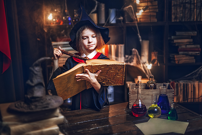 Kleine Hexe liest aus Zauberbuch und hält einen Zauberstab in den Händen
