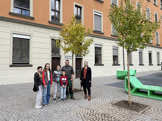 Zwei Mädchen und zwei Jungen stehen mit einem Stadtgärtner und Stadträtin Nathalie Koch im Gymnasiumhof bei den neu gepflanzten Bäumen.