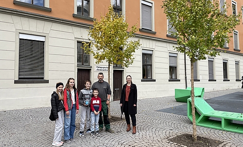 Zwei Mädchen und zwei Jungen stehen mit einem Stadtgärtner und Stadträtin Nathalie Koch im Gymnasiumhof bei den neu gepflanzten Bäumen.