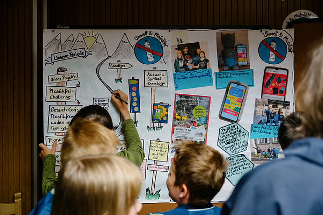 Kinder stehen vor einem bunt gestalteten Plakat, ein Kind zeichnet gerade auf dem Plakat.