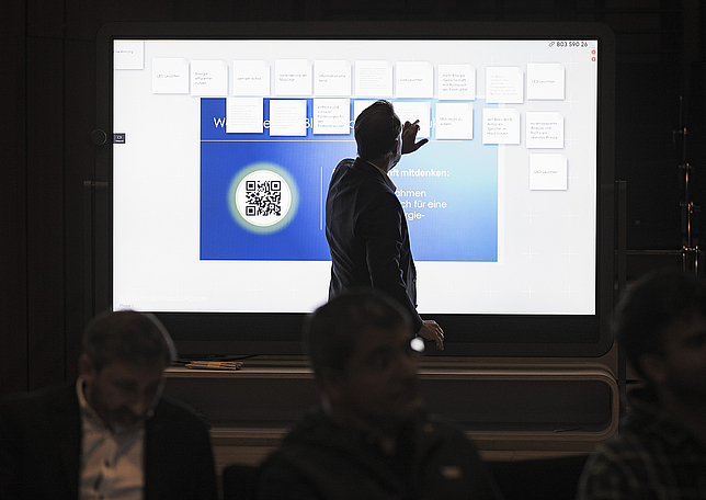 Digitalisierungskoordinator Alexander Maurer steht vor einem großen Bildschirm und tippt mit einem Finger darauf.