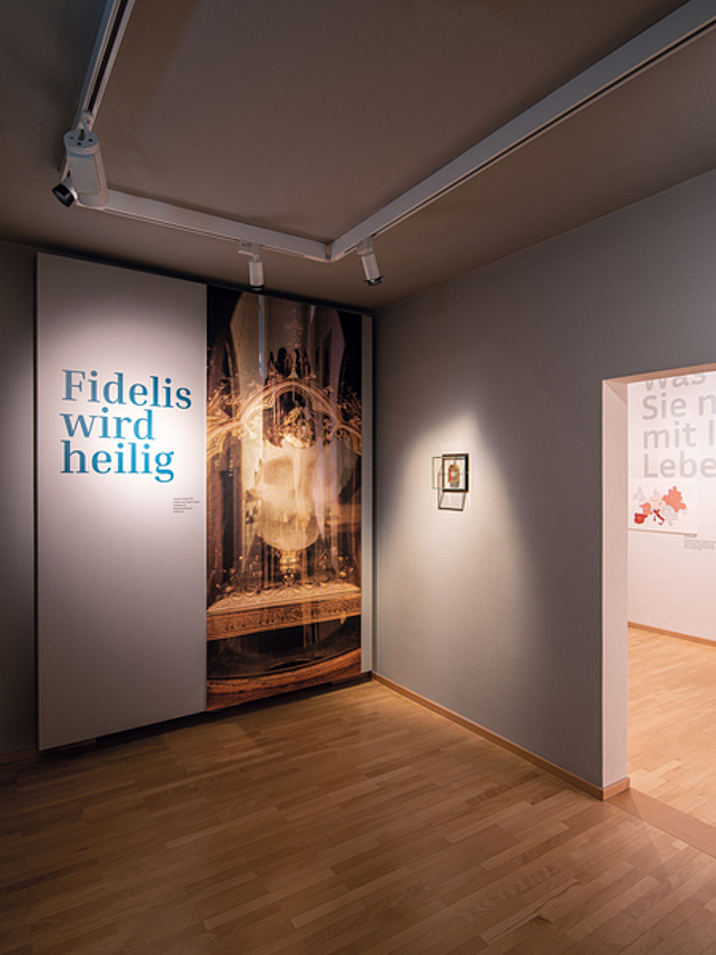 Ein Raum der Fidelis-Ausstellung im Palais Liechtenstein. An der Wand ist der Schriftzug "Fidelis wird heilig" zu lesen.