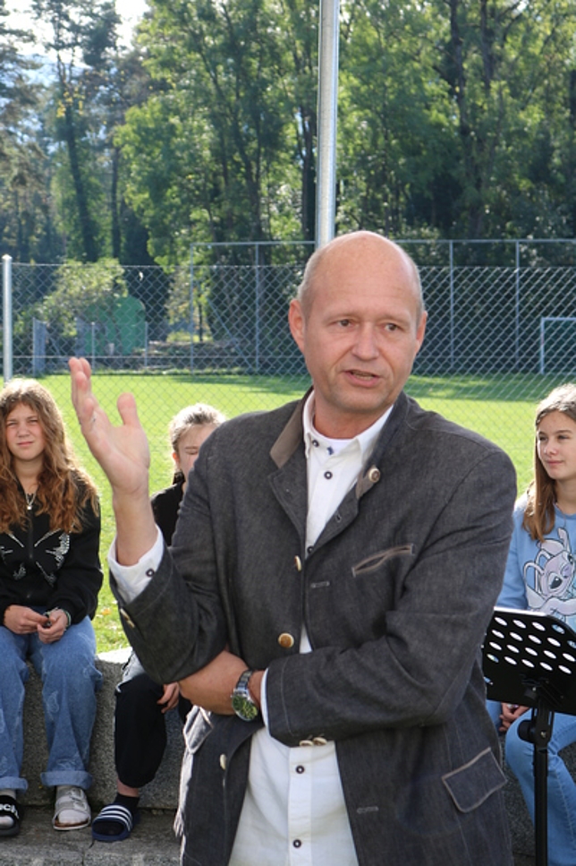 Direktor Gerold Zangerl spricht und gestikuliert, hinter ihm sitzen Kinder auf einer Stein-Mauer.