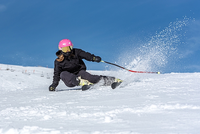Eine junge Frau fährt mit Ski einen mit schneebedeckten Berg hinunter, sie trägt einen pinken Ski-Helm.