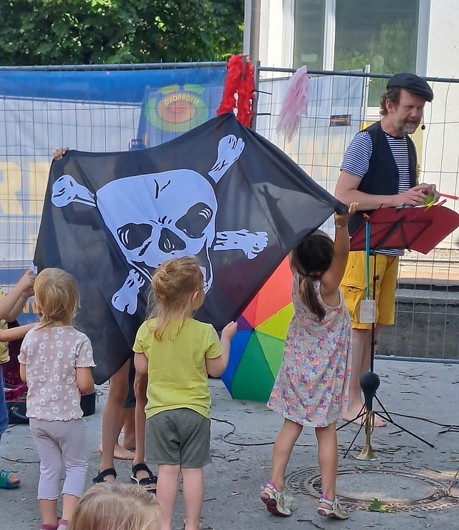 Ein Mann steht vorne und spricht in ein Mikrofon. Kinder stehen vor ihm und halten eine Flagge in die Luft.