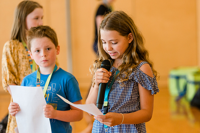 Ein Mädchen liest von einem Zettel ab und spricht in ein Mikrofon. Ein Junge steht daneben und blickt Richtung Kamera. 