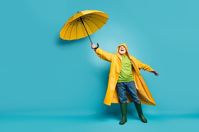 Ein älterer Herr im Regenmantel hält einen Regenschirm in der Hand und lacht. 