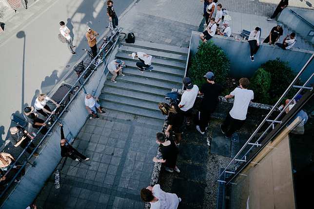 Eine Gruppe von Jugendlichen ist versammelt und schaut einem Skateboarder zu, wie er mit seinem Skateboard über eine Treppe springt.
