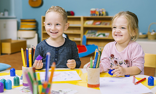 Zwei Kinder zeichnen im Kindergarten und lachen in die Kamera.