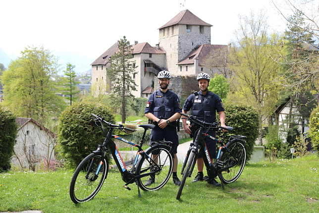 Zwei Polizisten tragen einen Fahrradhelm und ihre Uniform und stehen mit ihren Dienst-Fahrrädern auf einer Wiese. Im Hintergrund ist die Schattenburg.