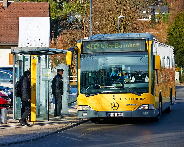 Ein Stadtbus hält an einer Haltestelle, an der drei Männer stehen. 