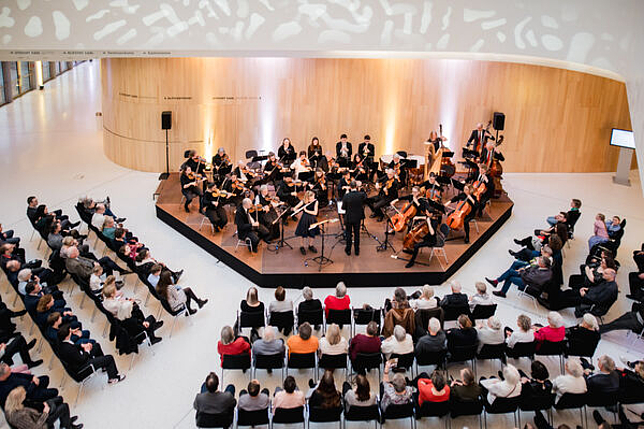 Orchester spielt im Atrium des Montforthauses, Besucherinnen und Besucher sitzen in gestuhlten Reihen vor dem Orchester..
