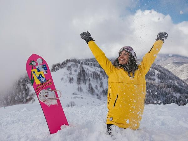 Ein Mädchen steht mit einer gelben Skijacke im Schnee und streckt die Hände nach oben. Neben ihr steckt ein pinkes Snowboard im Schnee.