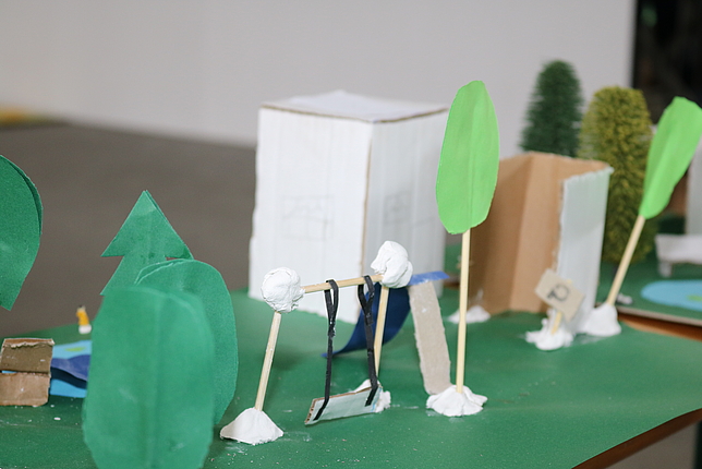 Auf einem grünen Blatt Papier sind durch Zahnstocher und Papier Bäume, eine Schaukel sowie eine Hütte in Miniaturansicht dargestellt