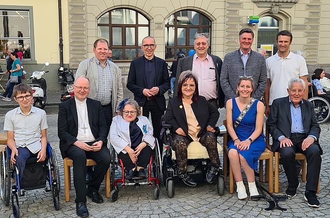 Fünf Männer stehen nebeneinander vor dem Dom in Feldkirch. Vor ihnen sitzen Männer und Frauen auf Stühlen oder in Rollstühlen. 