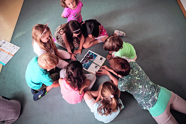 Eine Kindergartenpädagogin liegt mit einem Bilderbuch auf dem Boden. Mehrere Kinder sitzen im Kreis um sie herum. 