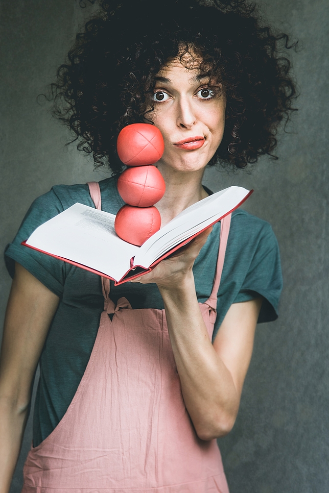 Eine Frau hält ein aufgeklapptes Buch mit einer Hand, auf dem aufgeklappten Buch liegen drei rote Bälle gestapelt übereinander.
