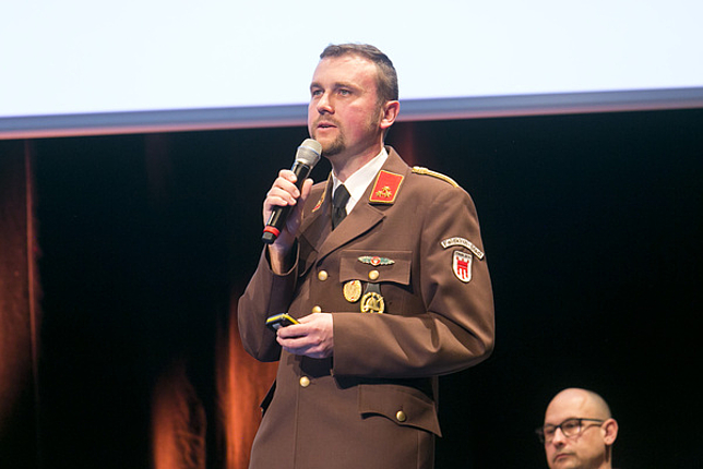 Kommandant der Feuerwehr Stadt Andreas Weiss steht auf der Bühne und spricht in ein Hand-Mikrofon.