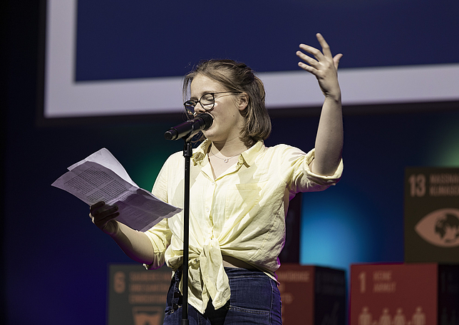 Ein junges Mädchen steht auf der Bühne und spricht in ein Mikrofon, das vor ihr steht. In einer Hand hält sie Zettel, von denen sie abliest. Die andere Hand hält sie nach oben.
