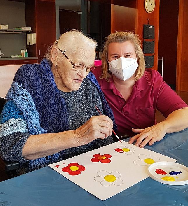 Eine Heim-Bewohnerin und eine Pflegerin malen Blumen auf ein Blatt Papier.