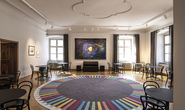 Raum im Palais Liechtenstein, ein großer runder Teppich mit bunten Streifen, um den Teppich herum stehen schwarze Tische und Stühle.