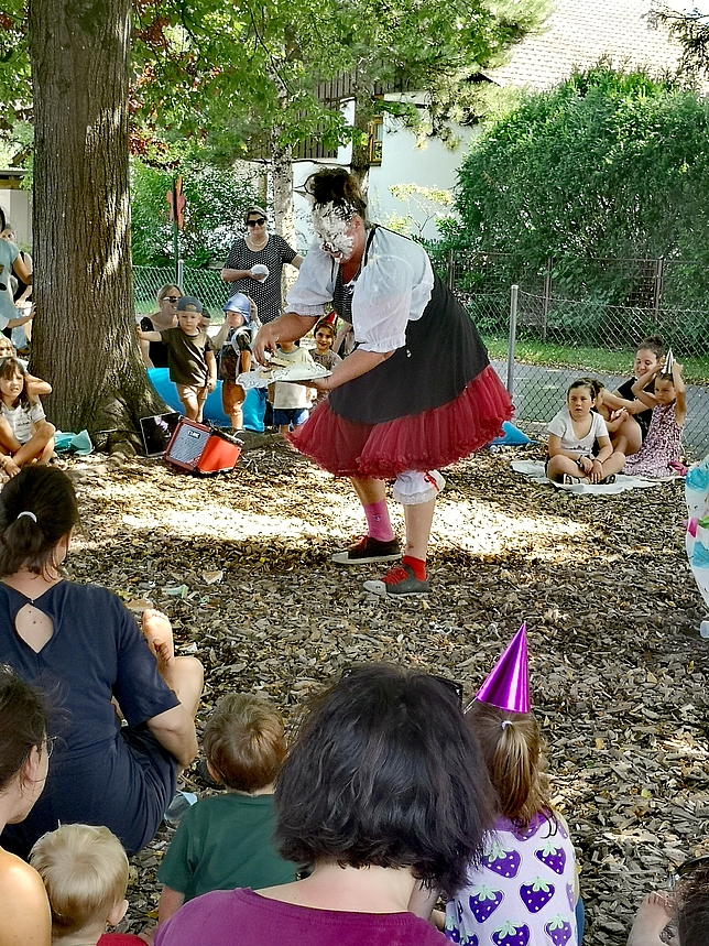 Eine Clownfrau steht unter einem Baum, um sie herum sitzen Kinder mit ihren Eltern. Die Clownfrau hat Torte im Gesicht.