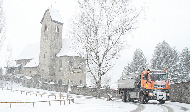 LKW der Stadt Feldkirch fährt auf schneebedeckter Straße, im Hintergrund ist eine Kirche zu sehen.