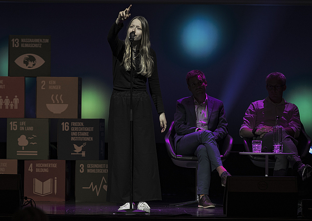 Ein junges Mädchen steht auf der Bühne und spricht in ein Mikrofon, dass vor ihr steht. Sie zeigt mit einer Hand nach oben, hinter ihr sitzen zwei Männer.