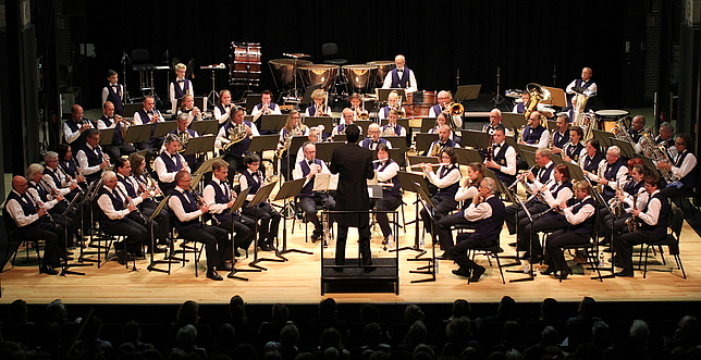 Ein Orchester sitzt im Halbkreis auf einer Bühne und spielt ihre Instrumente. Der Dirigent steht vorne vor dem Orchester.