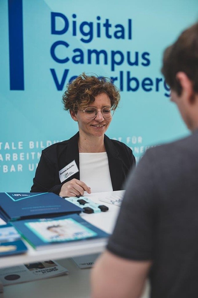 Eine Mitarbeiterin des Digital Campus Vorarlberg beim Informationsstand. Ein Besucher steht beim Informationsstand.